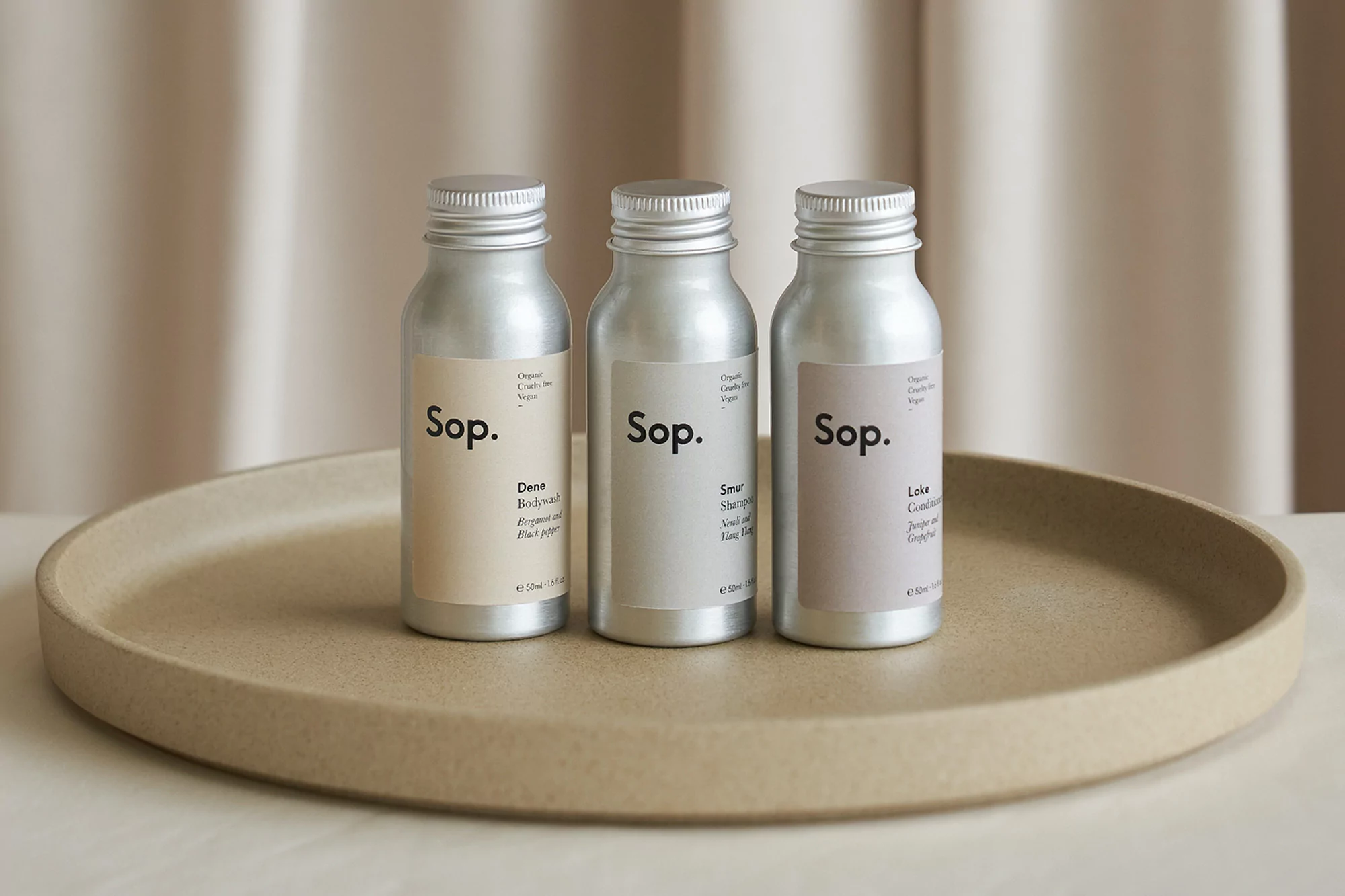 Three aluminum bottles of Sop. brand organic bodywash, shampoo, and conditioner sit on a round beige tray against a neutral curtain backdrop.