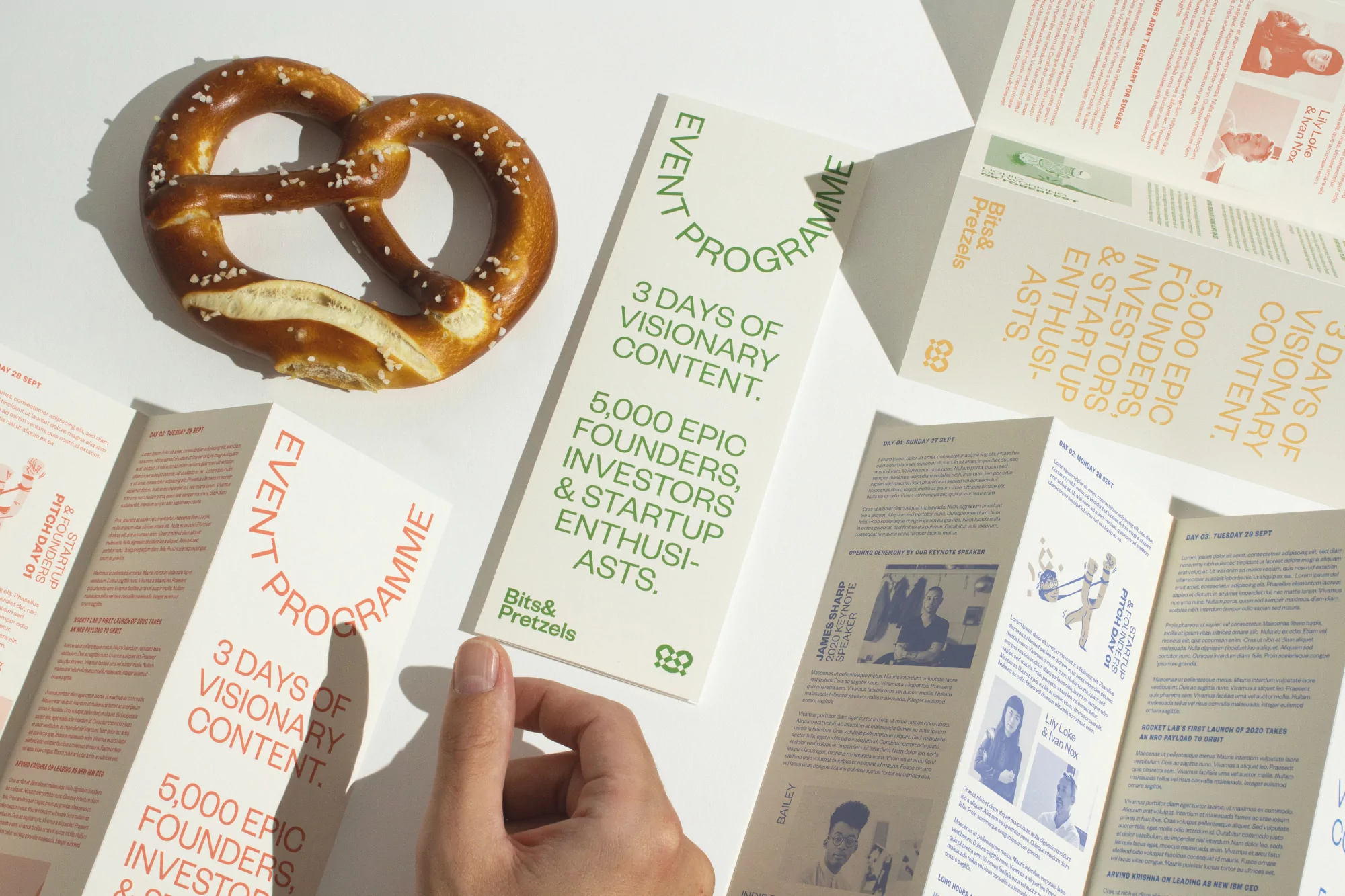 Overhead shot of a pretzel, hand, and several tri-fold brochures featuring event program information on a white surface with natural lighting.