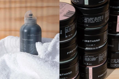 Firsthand Supply dark grey pump bottle nestled in a white towel with a stack of black pomade jars beside it.