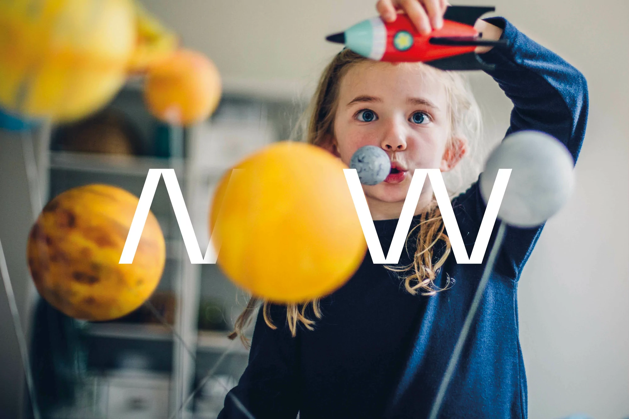 A young girl in a blue shirt holds a toy rocket and poses with a solar system model featuring yellow, gray, and orange planets.