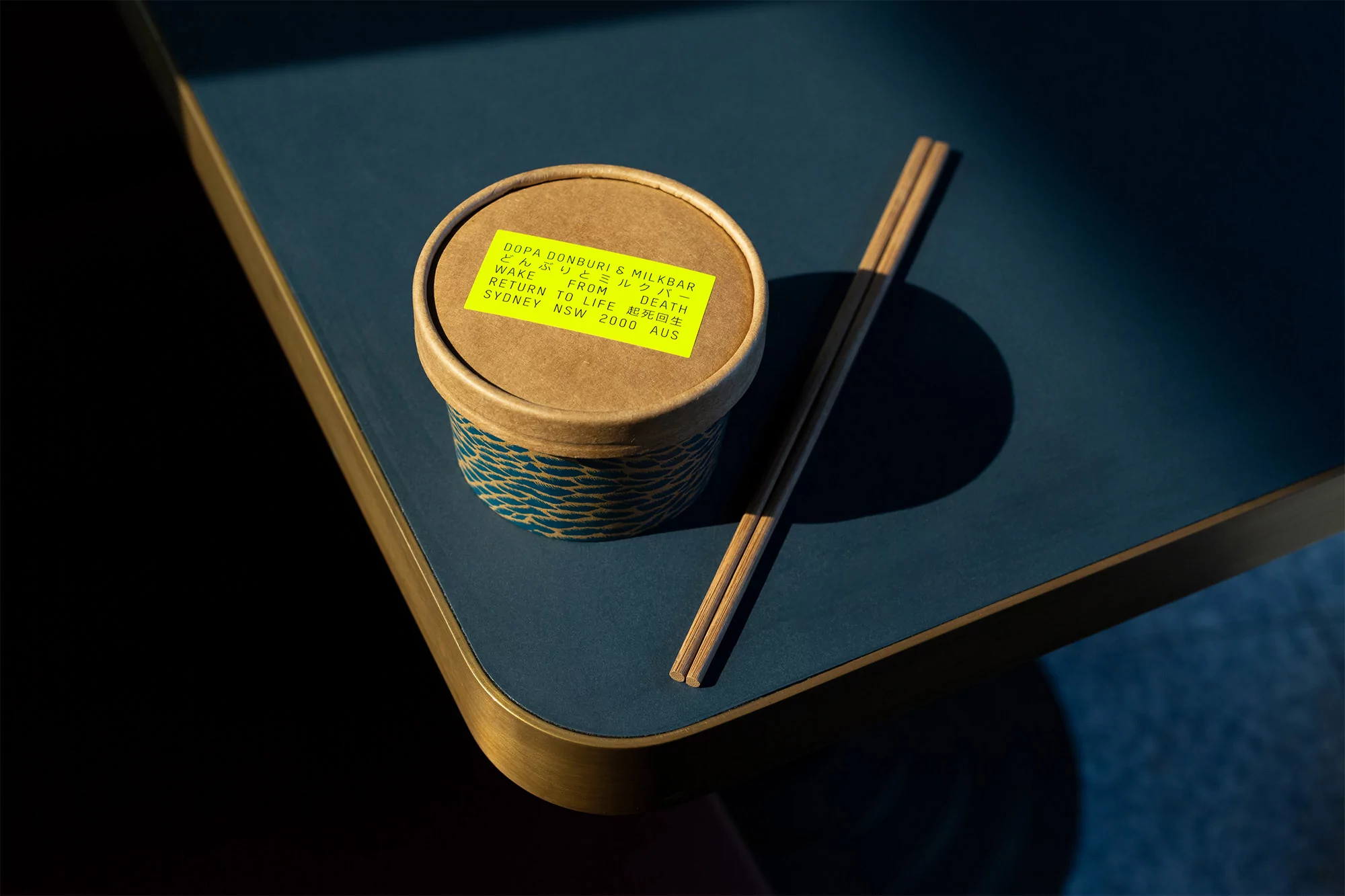 Close-up of a takeout container with chopsticks on a blue table. The container has a yellow label with black text. Food and Beverage.