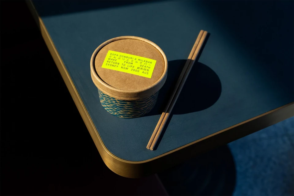 Close-up of a takeout container with chopsticks on a blue table. The container has a yellow label with black text. Food and Beverage.
