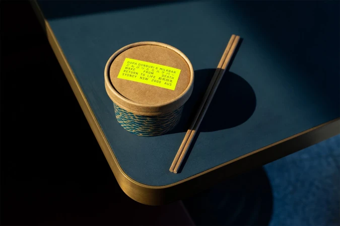 Close-up of a takeout container with chopsticks on a blue table. The container has a yellow label with black text. Food and Beverage.