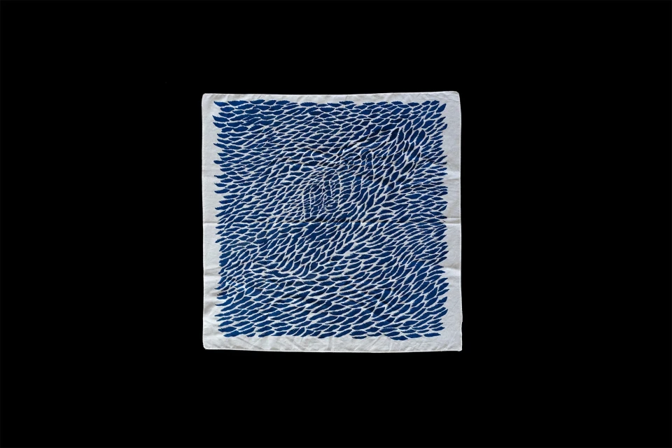 Square piece of white textile art with dense blue pattern resembling leaves, against a solid black background.