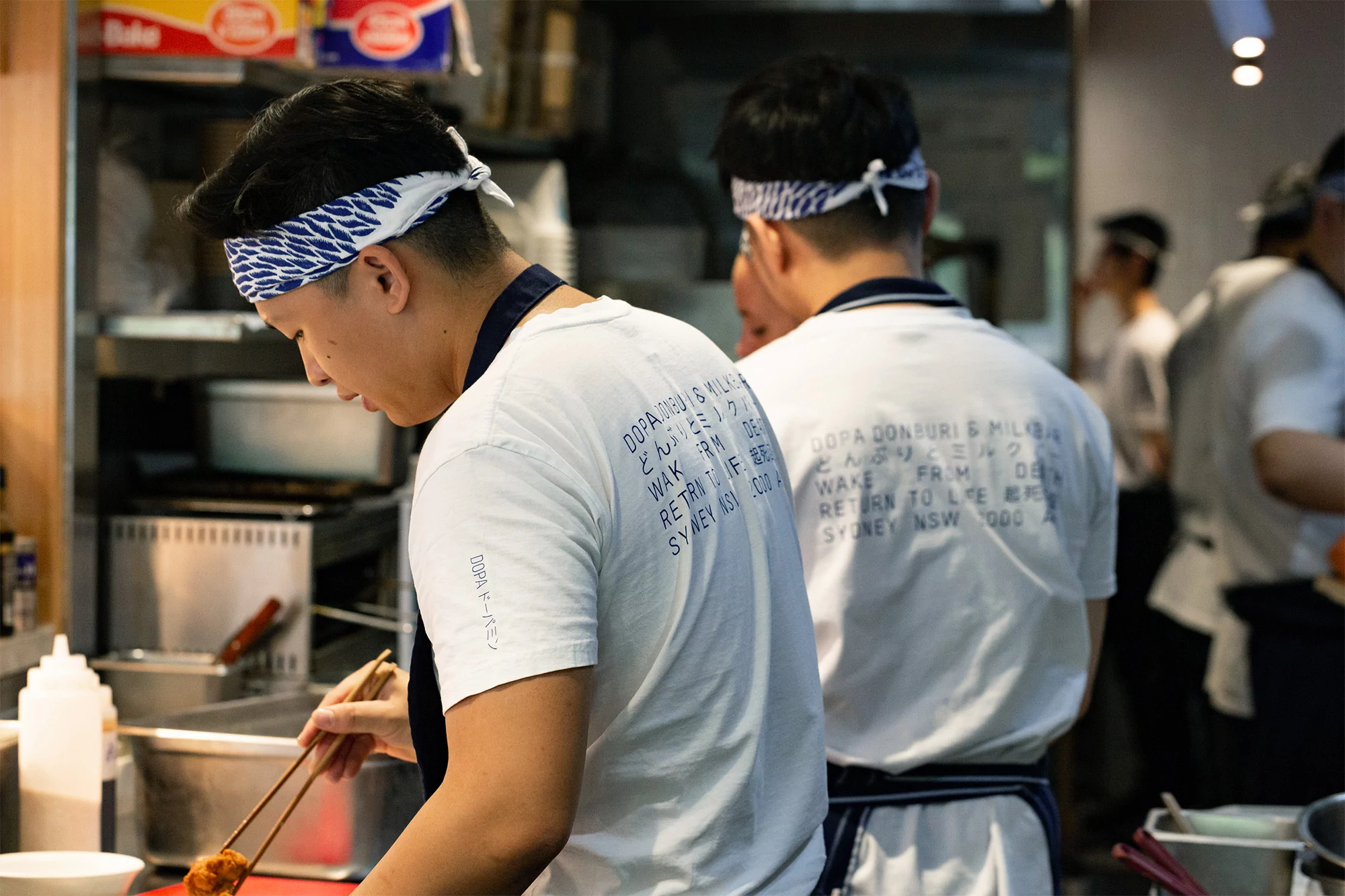 Chefs in white shirts and headbands working in a commercial kitchen with stainless steel appliances and cooking equipment visible.