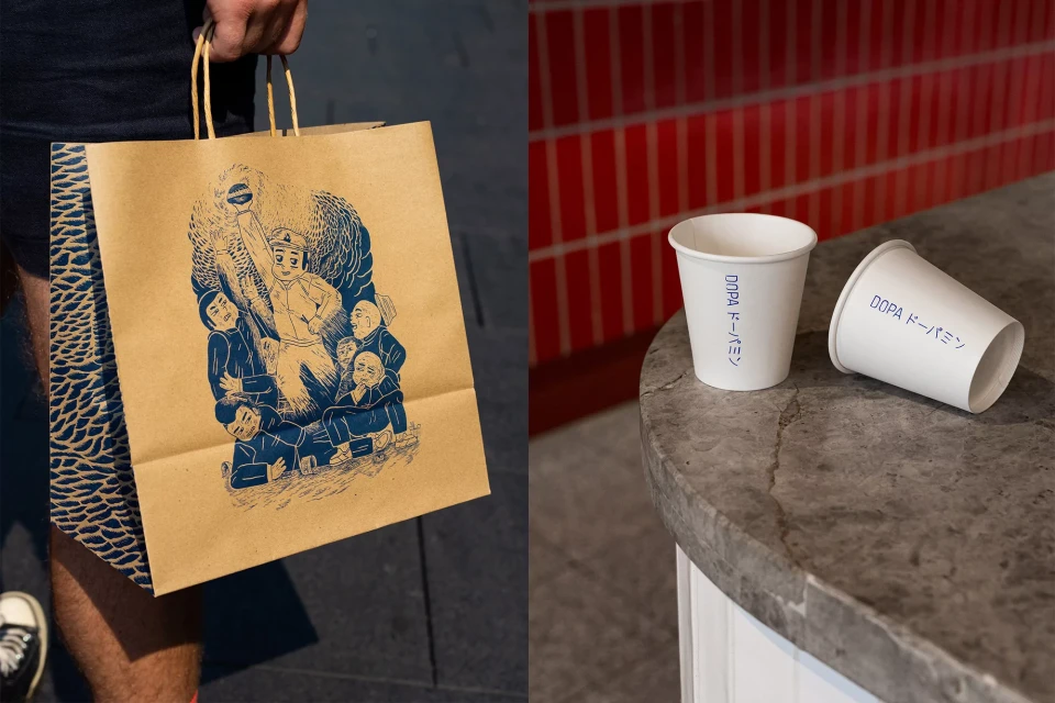 Person carrying a paper bag with an illustration of people, next to coffee cups on a marble counter with red brick tiles behind.