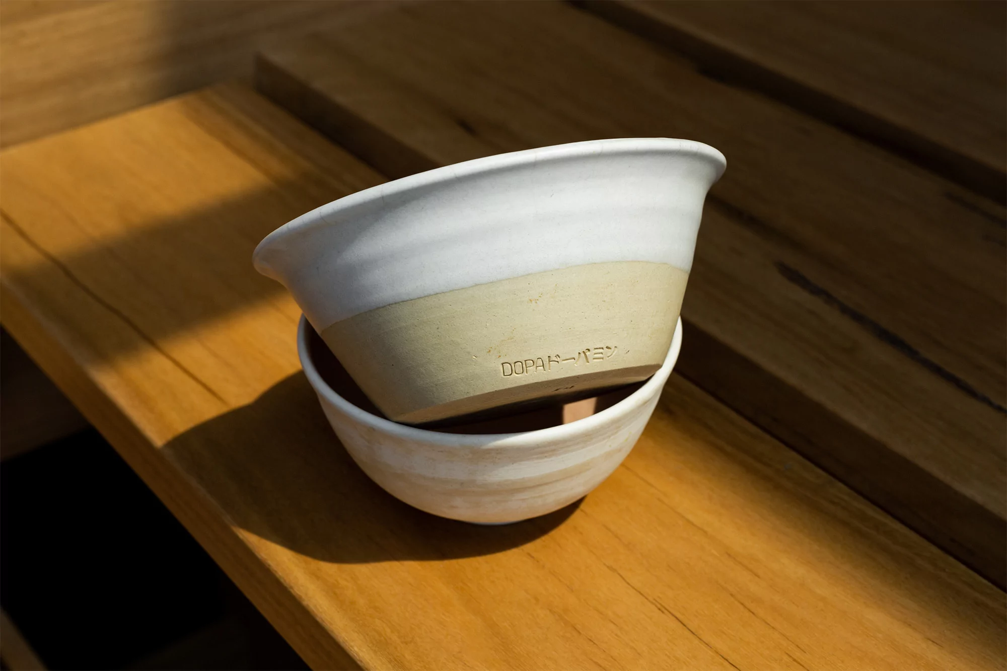 Two stacked ceramic bowls, the top one white and tan, sit on a wooden surface under natural light, showcasing their handmade aesthetic.