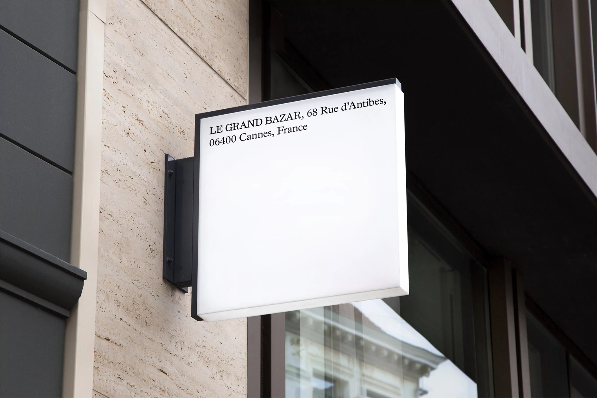 Square illuminated sign on a building facade, displaying 'LE GRAND BAZAR, 68 Rue d'Antibes, 06400 Cannes, France' in black text.