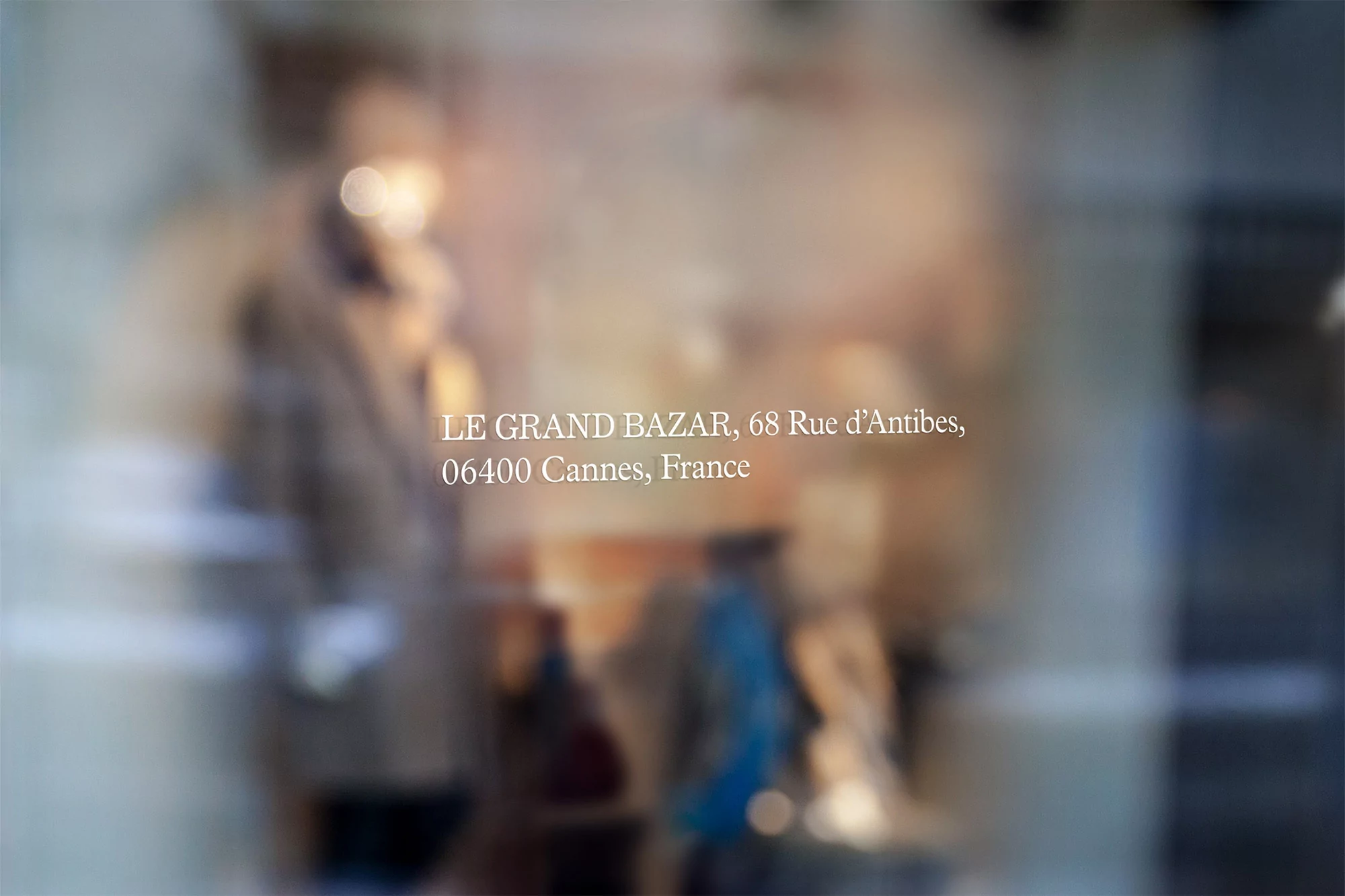 Storefront window with text 'LE GRAND BAZAR, 68 Rue d'Antibes' and blurred figures in background; captures a street scene in Cannes, France.