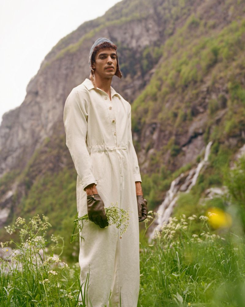 Man in off-white coveralls holds wildflowers in a green field against a mountain backdrop on an overcast day.