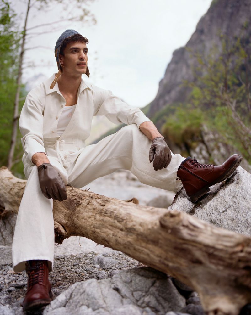A young man in a white outfit and hat sits on a log in a rocky, mountainous area wearing work boots and gloves in a daytime shot.