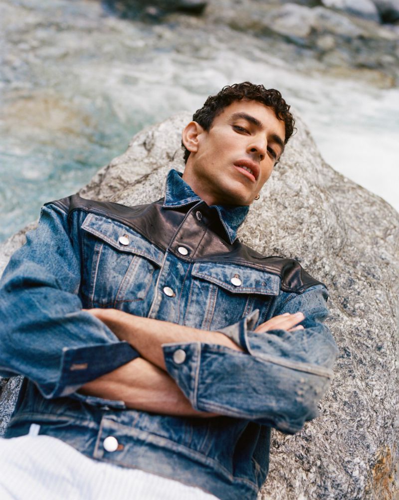 Portrait of a man in denim jacket posing outdoors, leaning against a rock with flowing water in the background, looking at the camera.