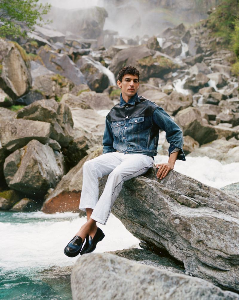 An adult male fashion model poses while sitting on a large rock in front of a cascading waterfall, wearing a denim jacket and white pants.