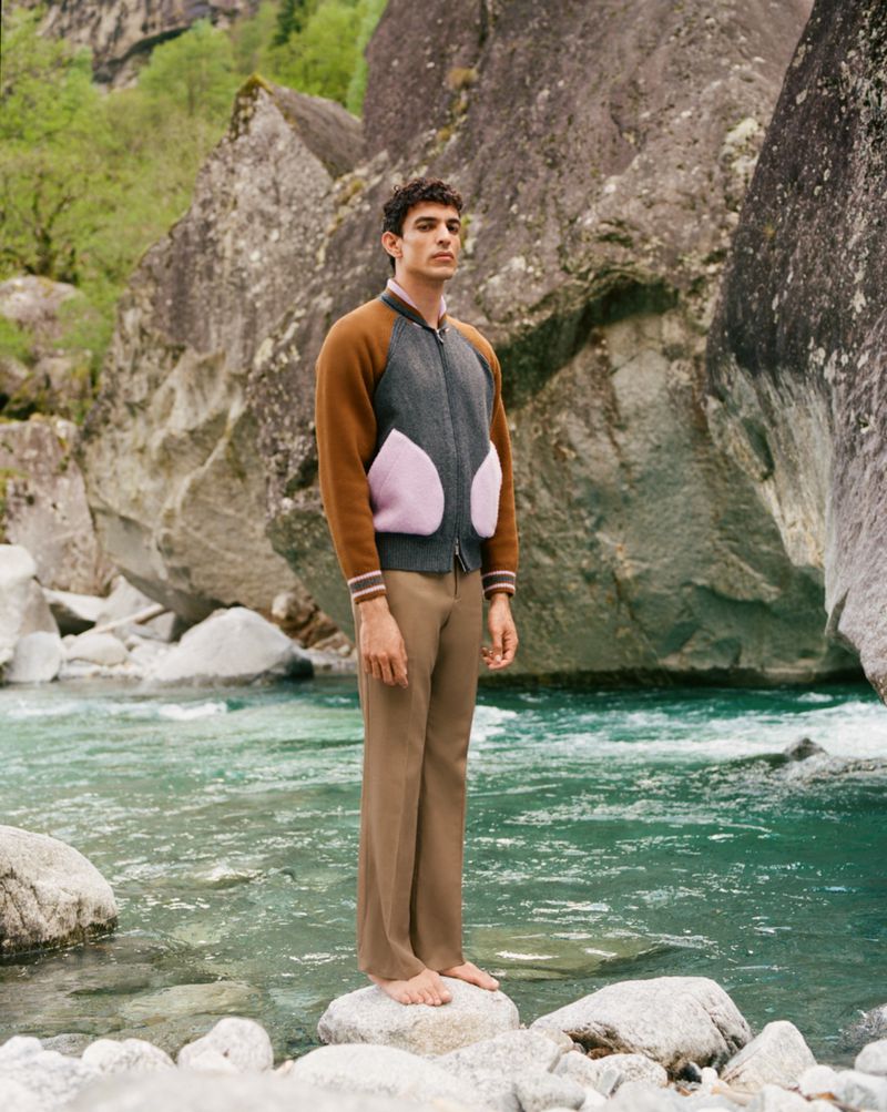 A barefoot man stands on rocks in a river, wearing a bomber jacket and tan pants, with large boulders and green trees in the background.