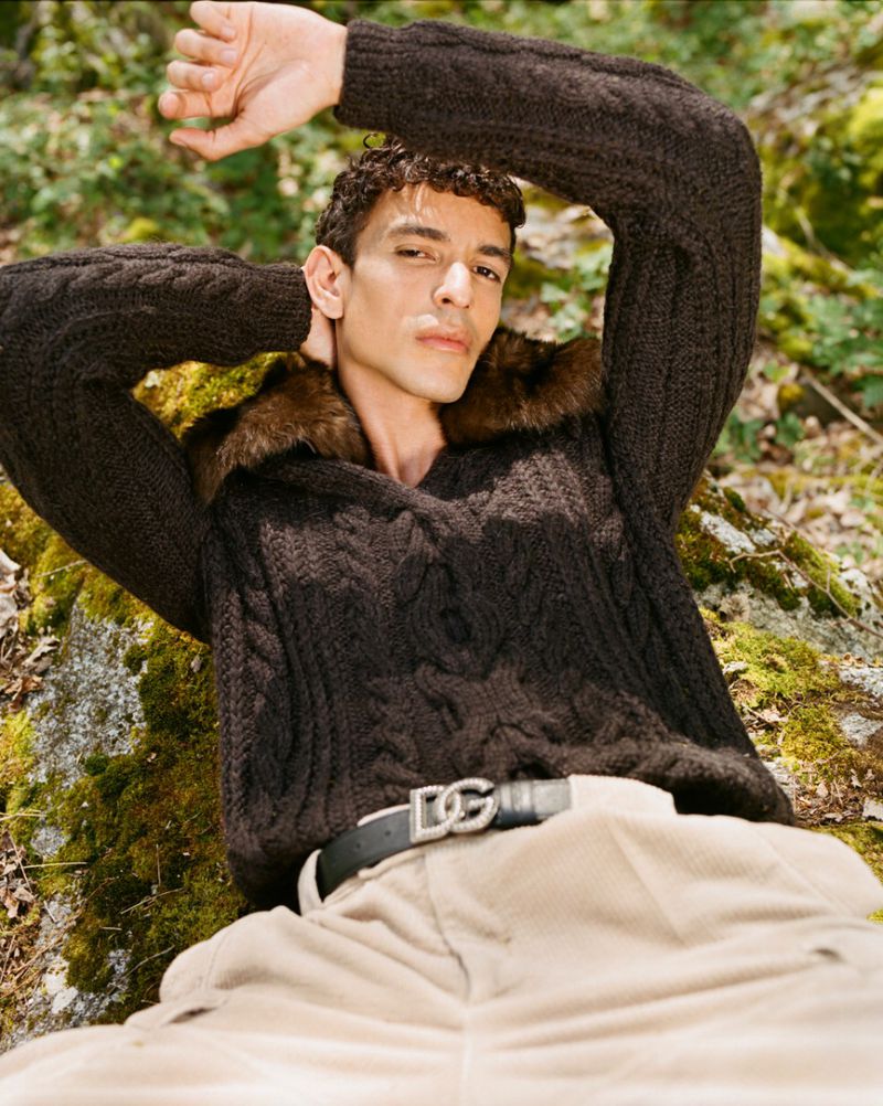 A young adult man poses lying down in a forest wearing a dark brown cable knit sweater, light beige trousers, and a black leather belt.