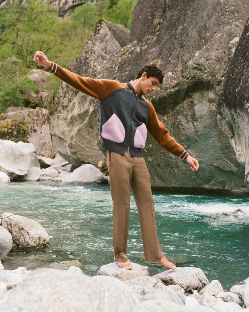Adult male balances barefoot on rocks in a flowing river, wearing tan trousers and a unique zip-up jacket in an outdoor mountain setting.