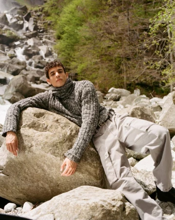 Man in grey sweater and light trousers rests on a rock in front of a waterfall and forest on a sunny day in an outdoor setting.