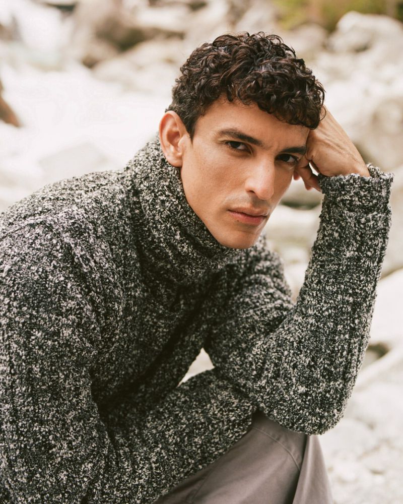 Close-up of a man with curly hair wearing a gray textured turtleneck sweater, gazing at the camera outdoors with rocky background.