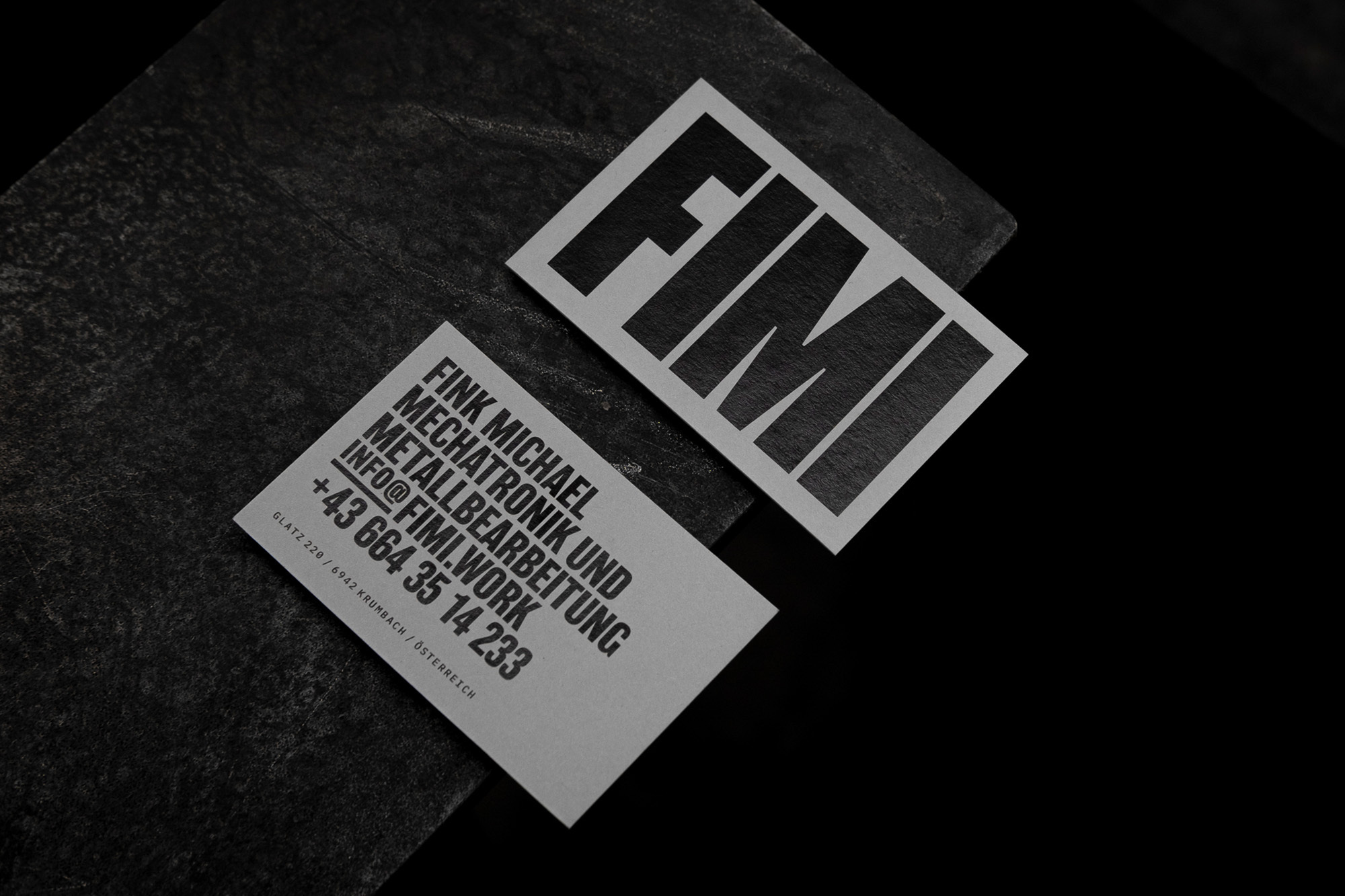 Two light grey business cards with black sans-serif typeface against a black background. One card is on a dark grey textured stone.