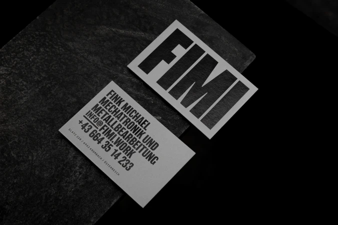 Two light grey business cards with black sans-serif typeface against a black background. One card is on a dark grey textured stone.