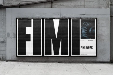 Three black and white posters spelling FIMI mounted on a grey urban wall above a concrete sidewalk.