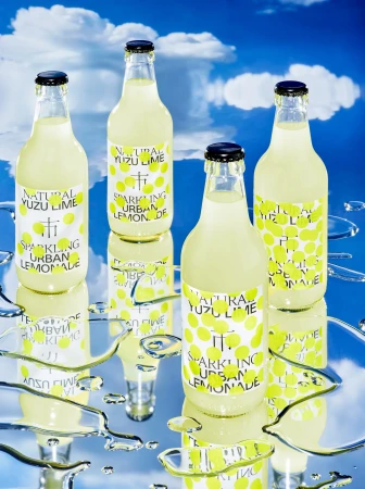 Four bottles of sparkling yuzu lime urban lemonade are arranged on a reflective surface against a backdrop of blue sky and clouds.