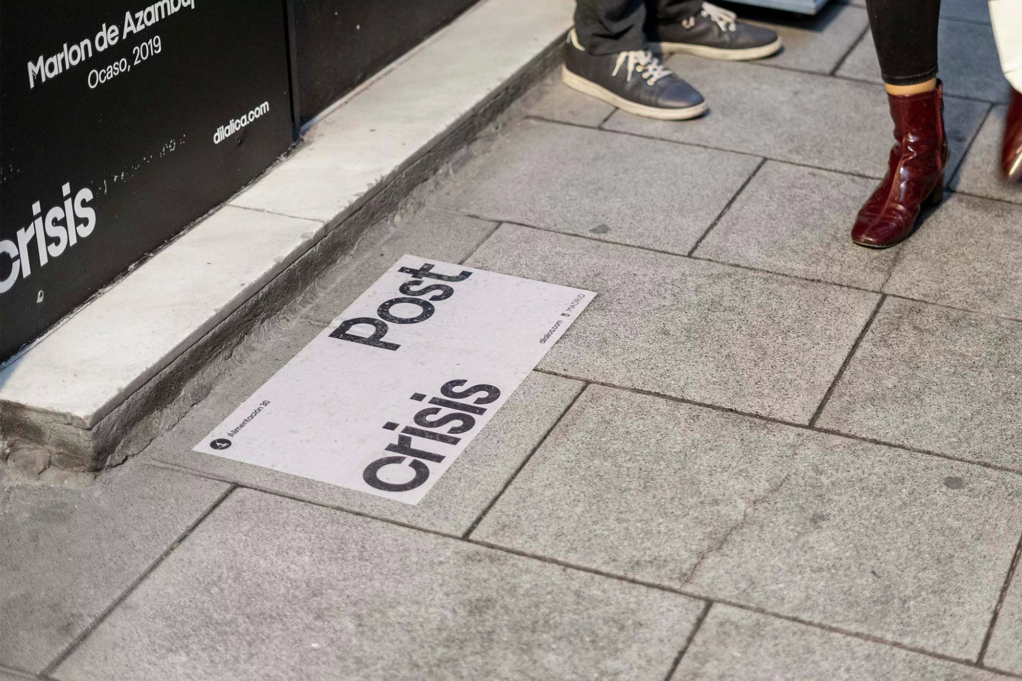 Sidewalk with text art installation reading 'Post Crisis,' featuring feet of passersby in casual shoes and dark trousers.