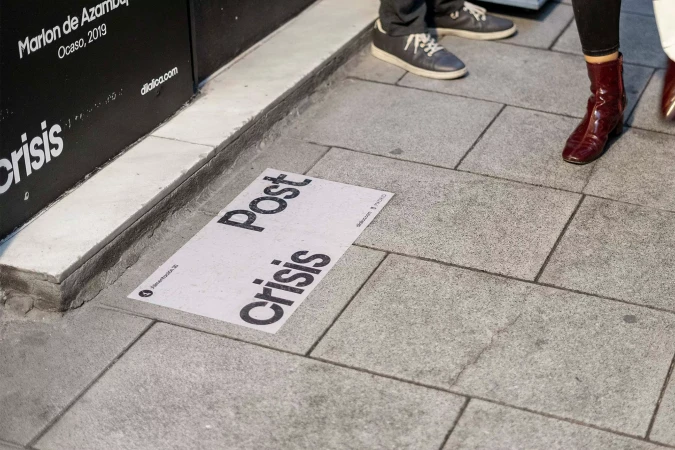 Sidewalk with text art installation reading 'Post Crisis,' featuring feet of passersby in casual shoes and dark trousers.