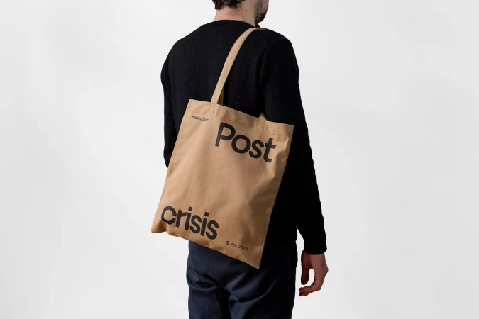 Adult man wearing a black long-sleeve shirt carries a tan tote bag with black text reading 'Post Crisis' in a studio setting.