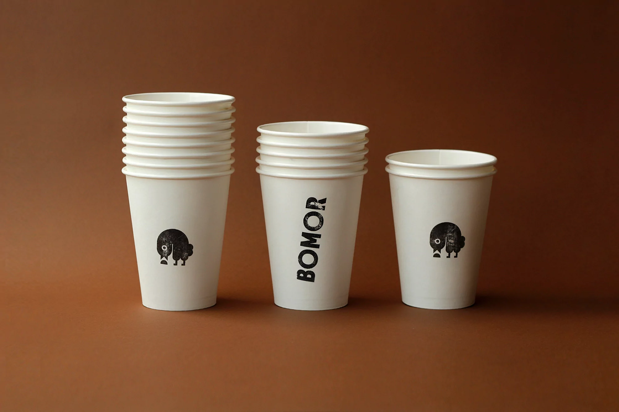 Three stacks of white paper cups sit on a brown surface, each featuring a black logo or text, creating a minimalist studio product shot.