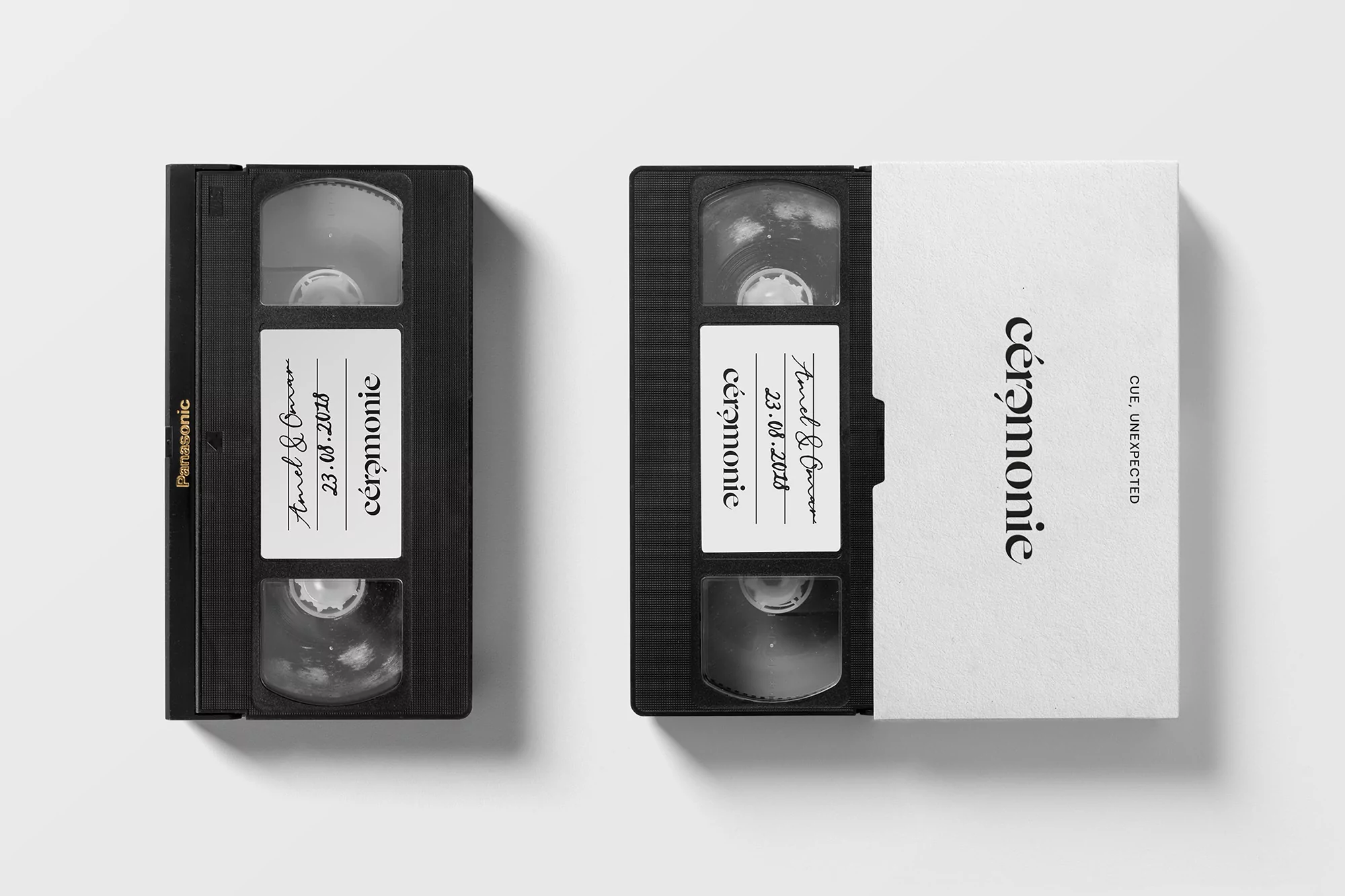 Two black VHS tapes with labels rest on a white surface in a studio setting. The tapes feature handwritten and printed text.