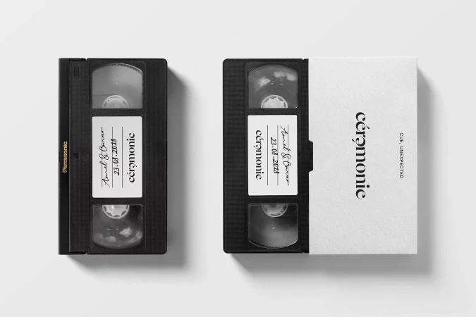 Two black VHS tapes with labels rest on a white surface in a studio setting. The tapes feature handwritten and printed text.