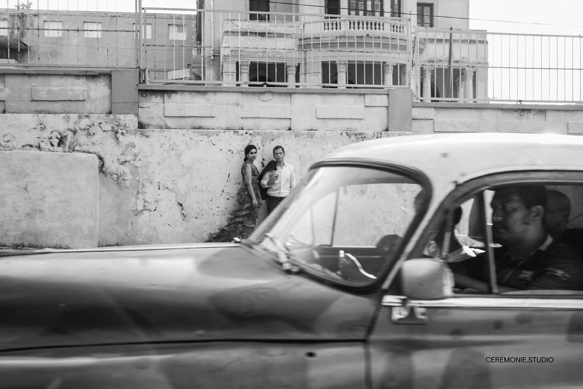 Black and white photograph of a couple standing near a textured wall while a classic car drives by on a city street.