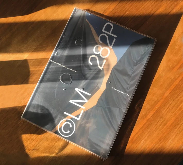 Still life photo of a book cover in its plastic wrap on a wooden surface, displaying gray, black, and peach hues in sunlight.