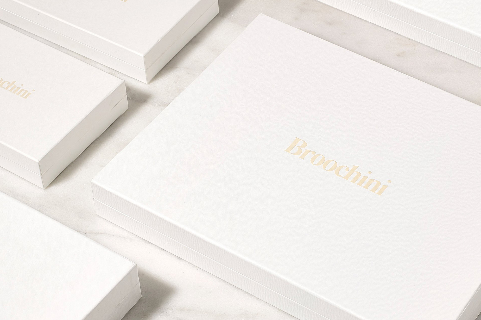 Close-up of a stack of elegant white square boxes with the brand name Broochini in gold lettering, stacked on a marbled surface.