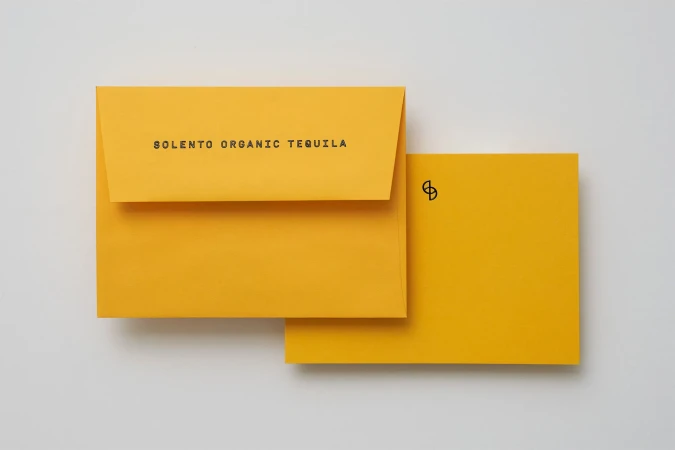 Still life shot of yellow envelope and card with SOLENTO ORGANIC TEQUILA text, against white backdrop in a studio setting.