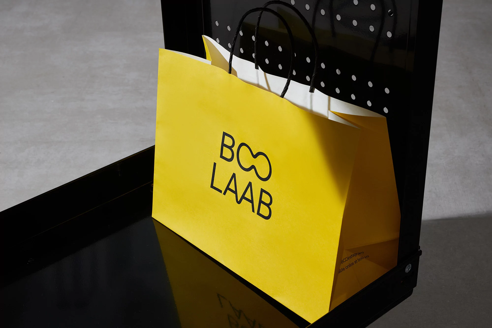 A yellow shopping bag with a black logo sits on a black surface against a gray backdrop, providing a vibrant yet minimalist composition.