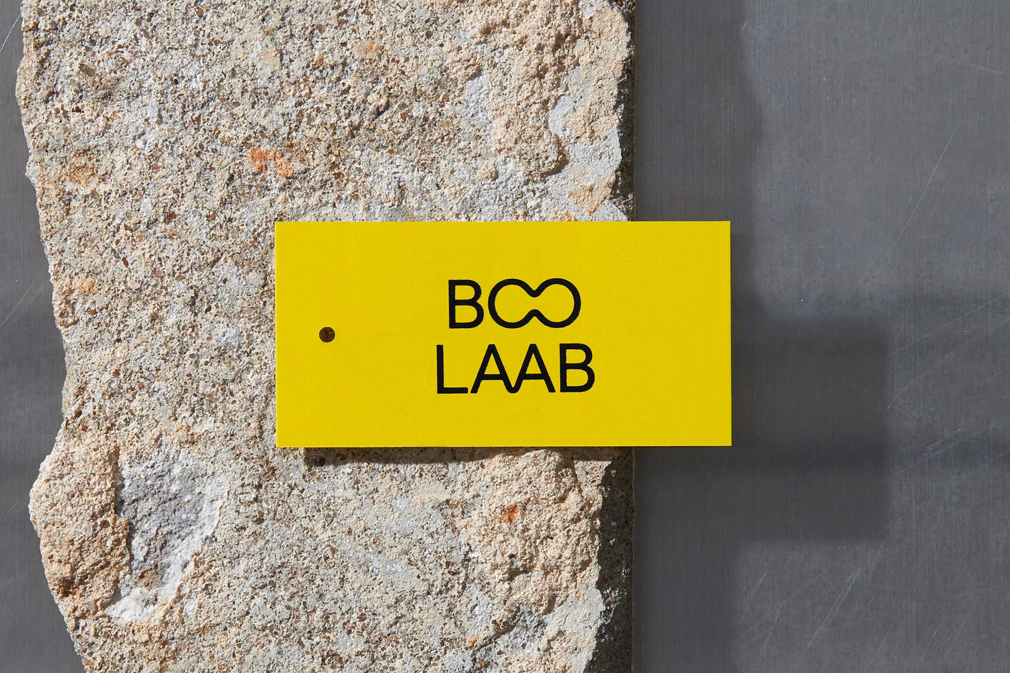 Yellow tag with black logo and text rests on a textured concrete block against a smooth grey surface under bright studio lighting.