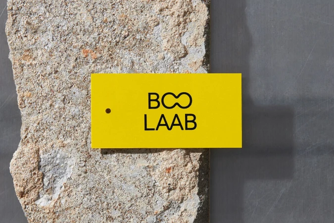 Yellow tag with black logo and text rests on a textured concrete block against a smooth grey surface under bright studio lighting.