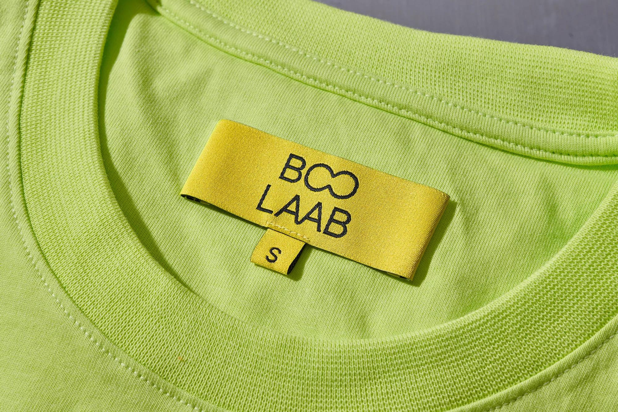 Close-up of a light green T-shirt with a yellow label featuring the brand 'BOO LAAB' and a size 'S', highlighting fashion details.