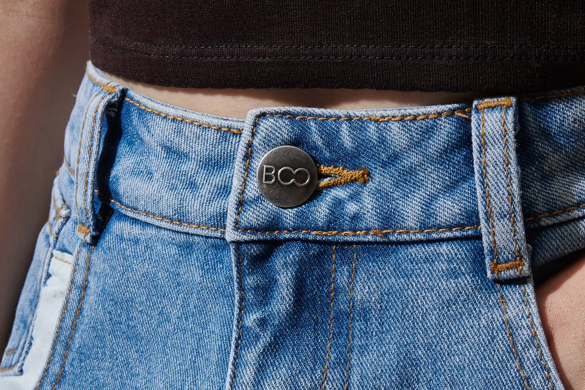 Close-up of light blue denim shorts with a button closure and brown threading, worn with a dark brown top, outdoor setting.