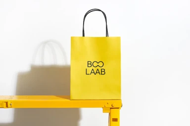 A bright yellow shopping bag with black handles and branding resting on a yellow surface against a plain white wall