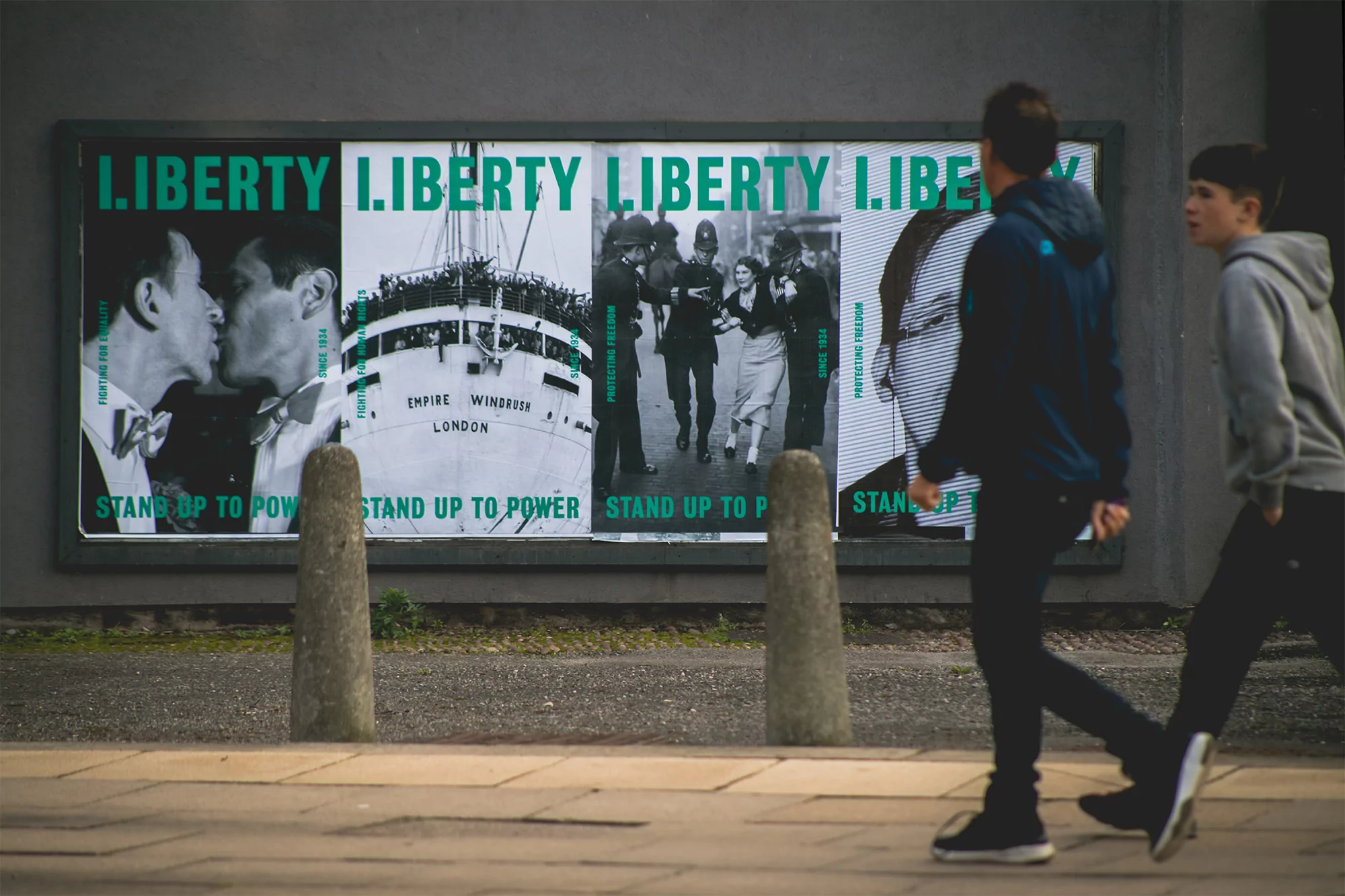 Two pedestrians walk past a display of four black and white posters on a gray wall, advertising freedom and justice.