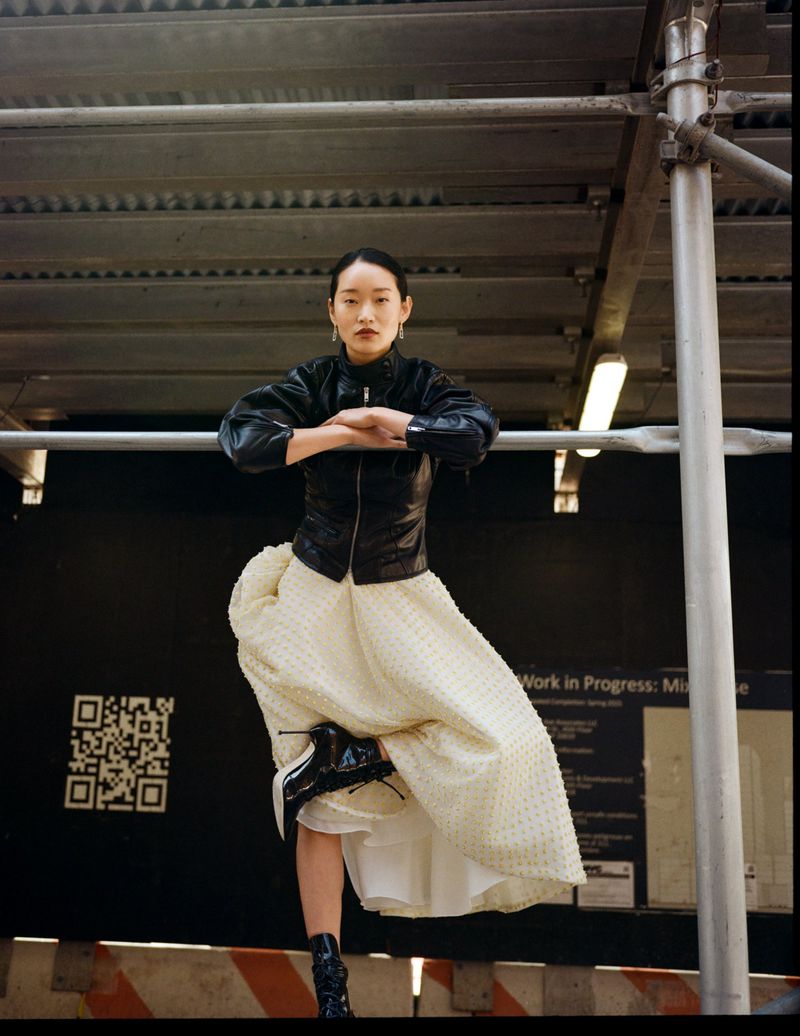 Young woman in a black jacket and white dress posing on a scaffolding in an urban setting, creating a unique fashion editorial look.