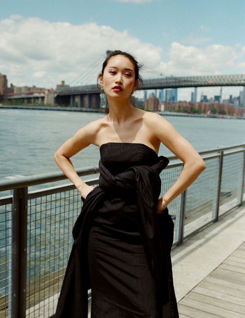 Asian female model poses in a black strapless dress on a boardwalk with a city and bridge in the background on a sunny day.