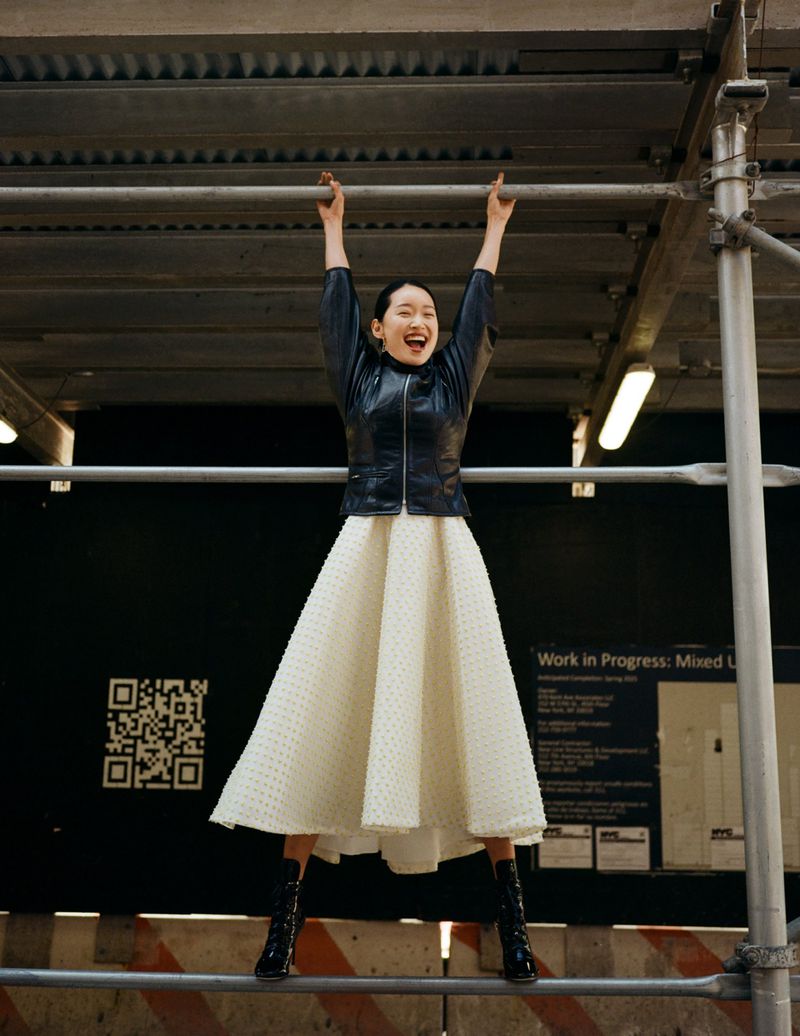An Asian woman with a wide smile stands on a scaffolding, wearing a black leather jacket, cream-colored dress, and black boots.