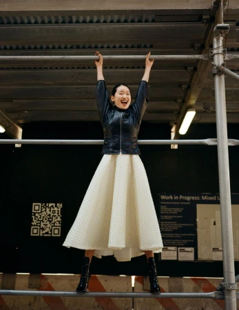 An Asian woman with a wide smile stands on a scaffolding, wearing a black leather jacket, cream-colored dress, and black boots.