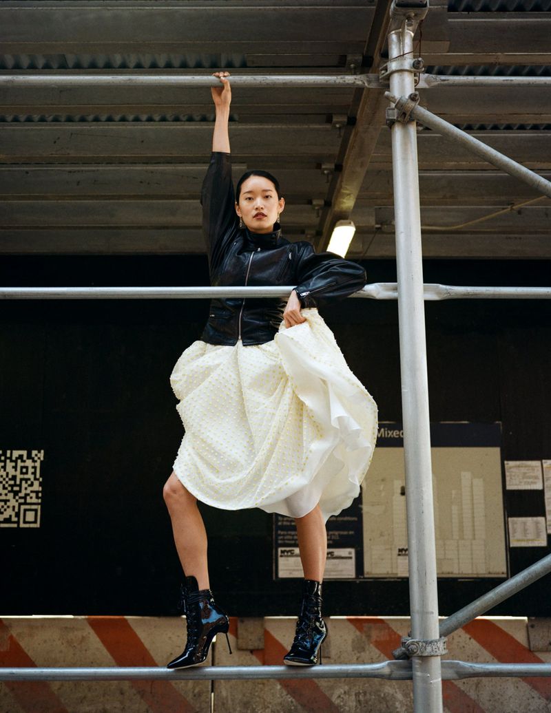 An Asian woman strikes a pose on scaffolding wearing a black jacket, full white skirt, and black heeled boots in an urban outdoor setting.