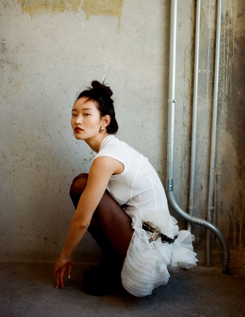 Asian woman in a white dress crouching in an indoor urban environment, posing with an interesting background with metal pipes.