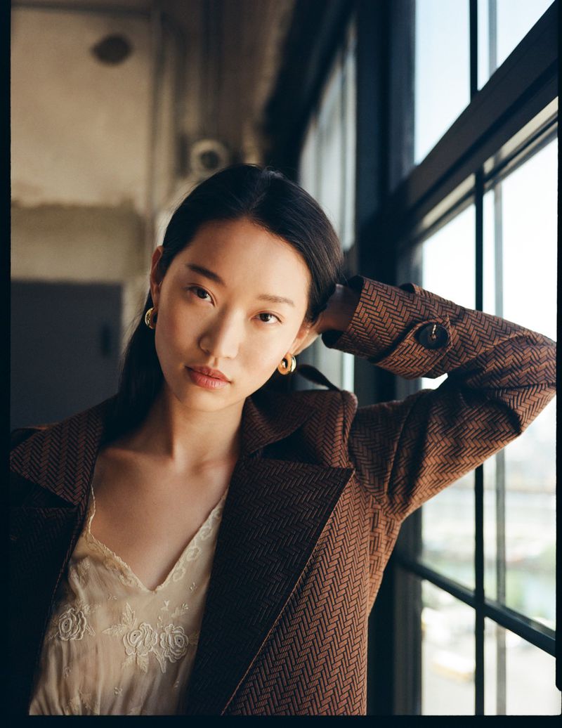 Asian woman poses near window in herringbone blazer and lace top, captured in a medium shot with soft, natural light indoors.